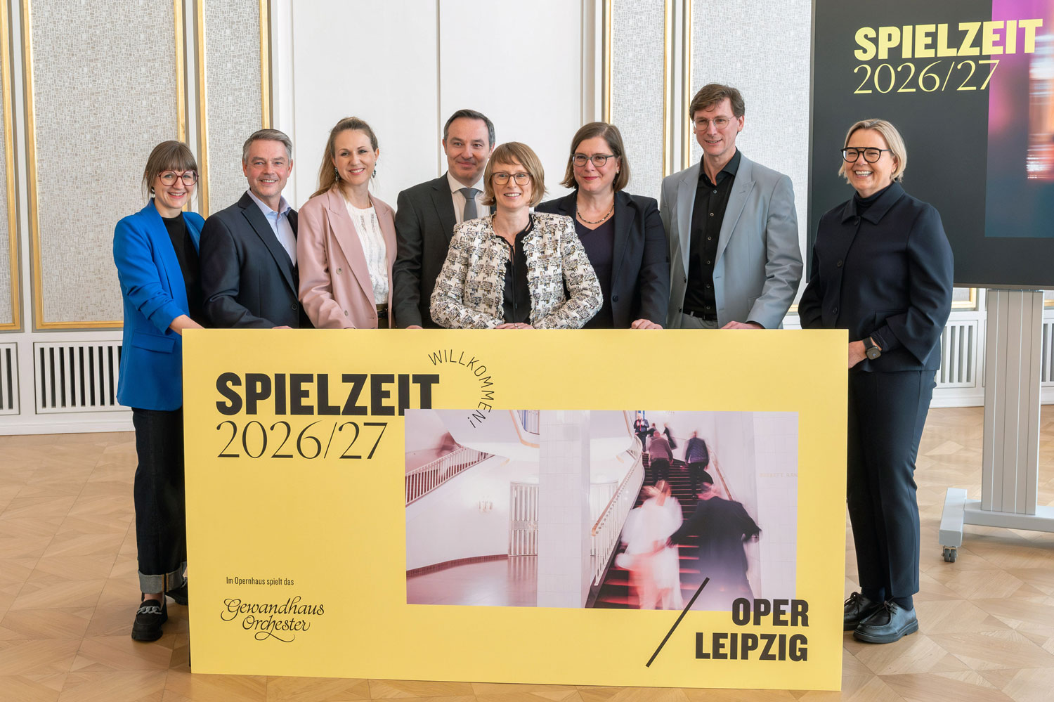 Acht Personen stehen im Foyer der Oper Leipzig hinter einem Tresen mit der Aufschrift Spielzeit 2026/27